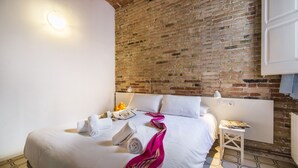 Una camera d'albergo con un muro in mattoni, un letto rifatto con lenzuola bianche, un comodino con una lampada e un pavimento piastrellato.