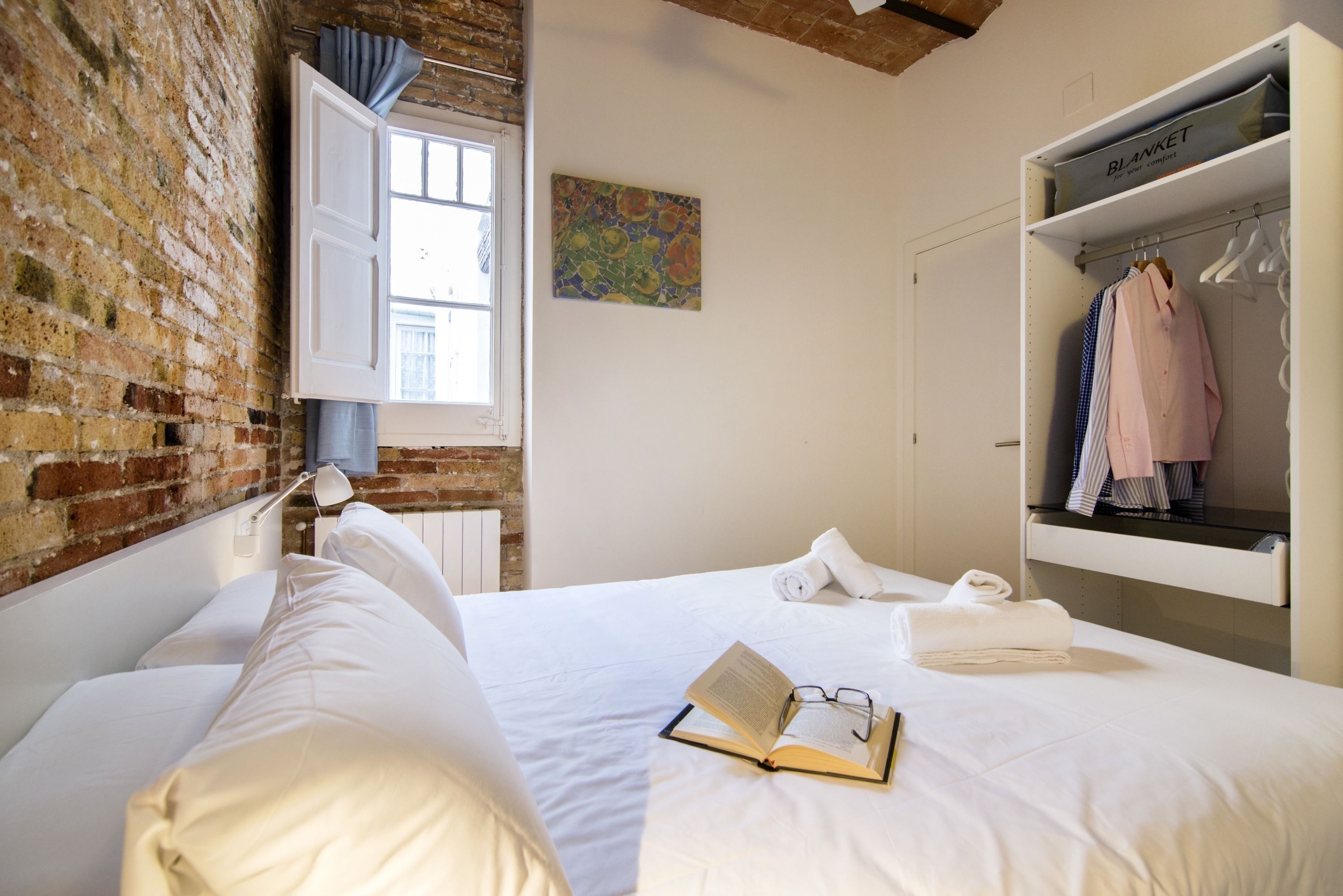 A bedroom with a brick wall, a window with curtains, a bed with white linens, a bedside table with a lamp, a bookshelf with clothes, and a painting on the wall.