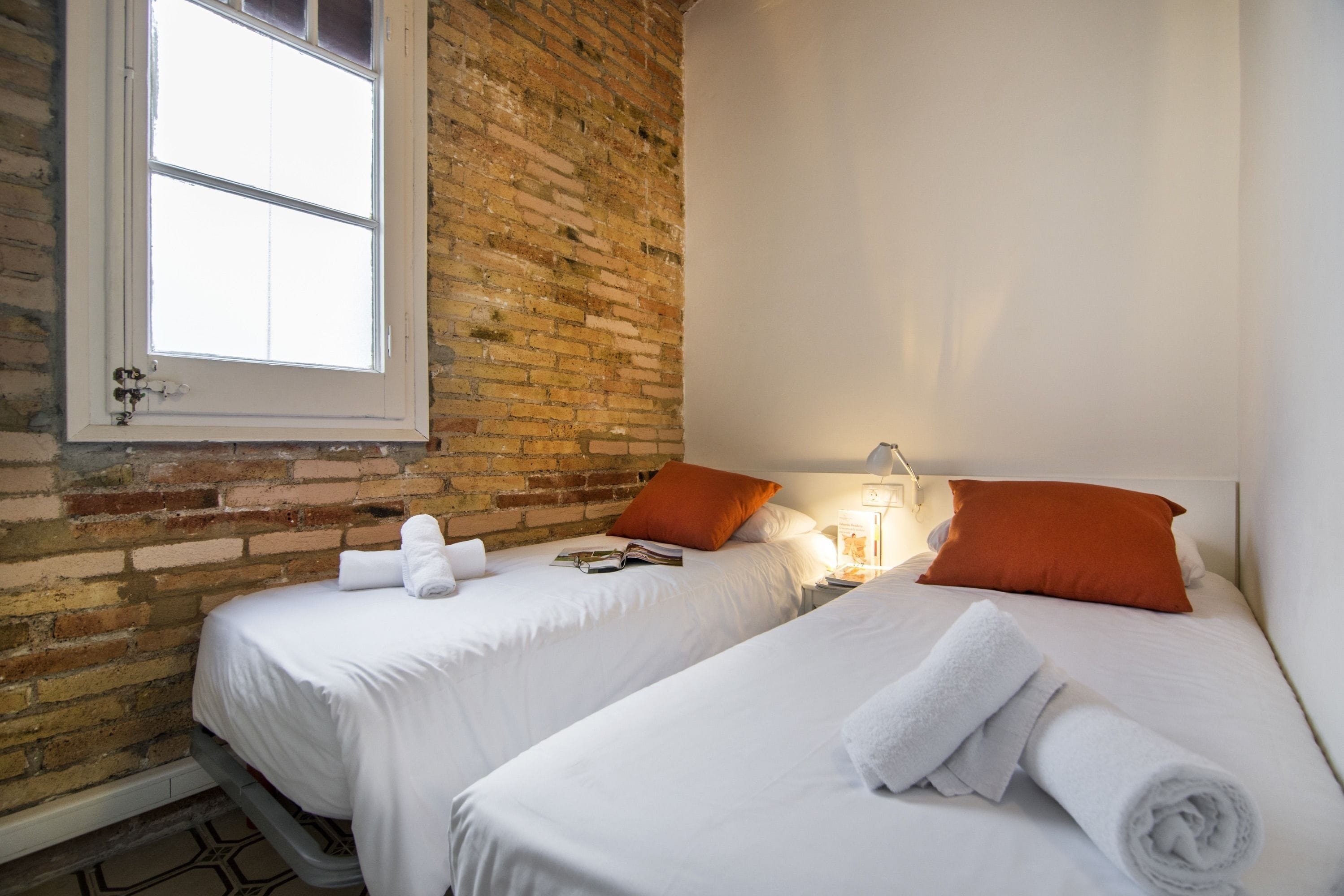 Una habitación con dos camas, ropa de cama blanca, un muro de ladrillo y una ventana.