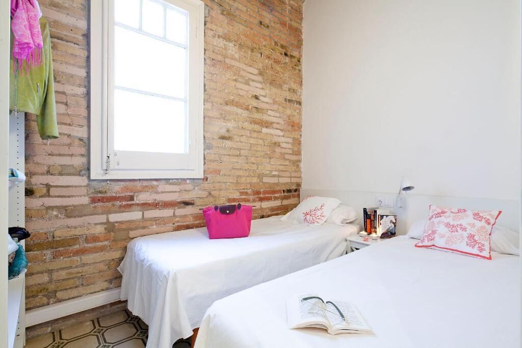 Una habitación con dos camas, una pared de ladrillo, una ventana y una mesita de noche con libros y una lámpara.