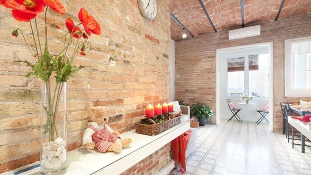 Una stanza con parete in mattoni, una sveglia, un vaso con fiori rossi, un orsacchiotto di peluche e un camino con candele.