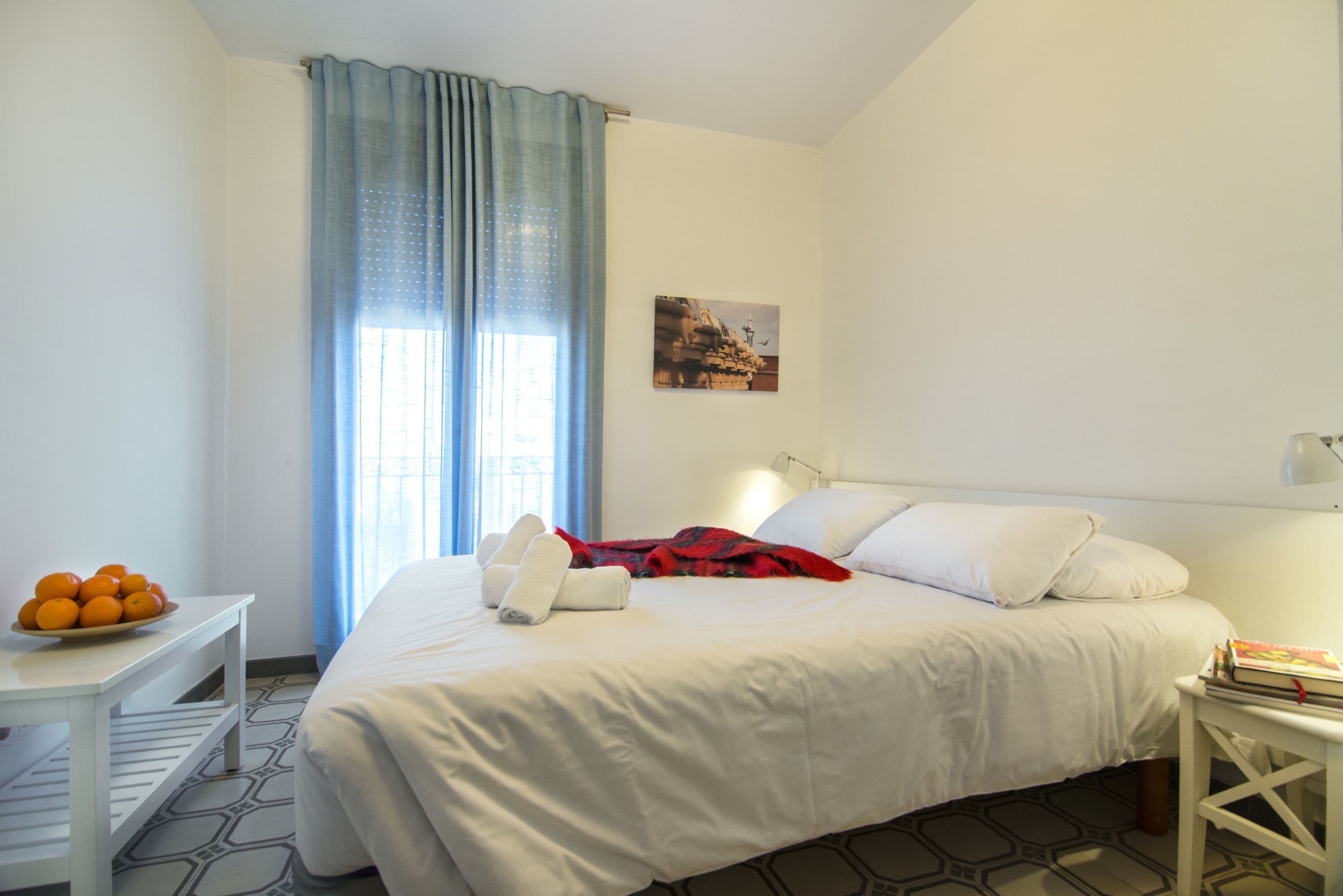 Una camera d'albergo con un letto, un comodino con una ciotola di arance, una lampada e un quadro appeso al muro.