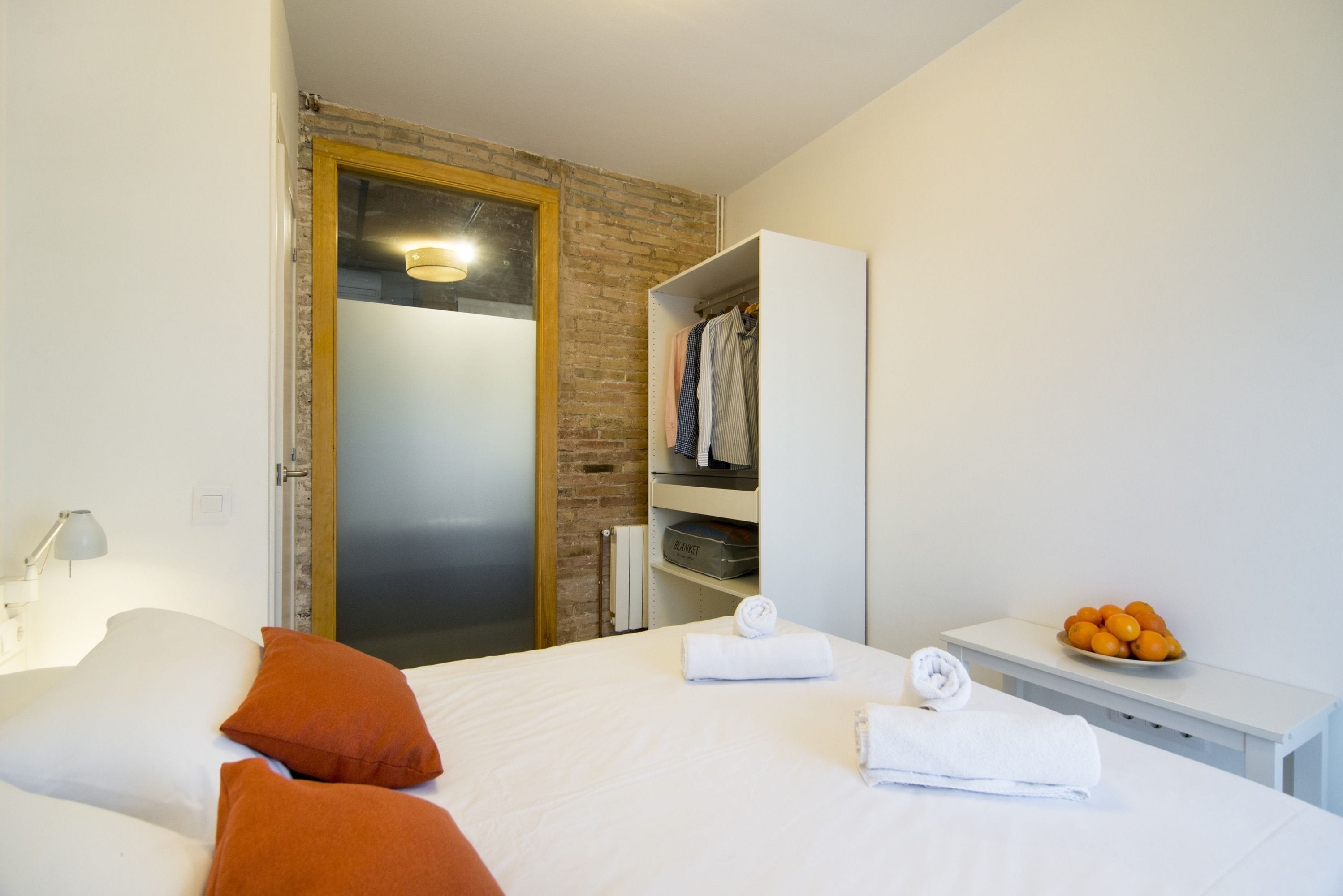 A hotel room with a bed, a wardrobe, a bedside table with a bowl of fruit, and a towel.