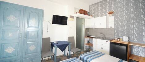 A compact hotel room with a kitchenette, a dining area with a table and chairs, a wardrobe, and a bed with patterned bedding.