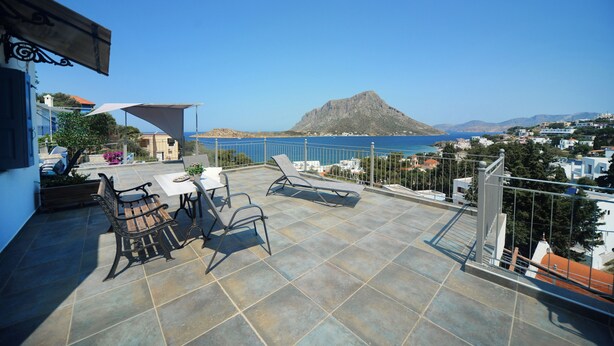 A terrace with a table and chairs, a sunshade, and a view of the sea and a mountain. - Panorama Studios & Apartments (Kalymnos)