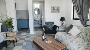 A living room with a checkered floor, a sofa, a coffee table with a bottle and bread, a chair, a lamp, and a painting on the wall. - Panorama Studios & Apartments (Kalymnos)