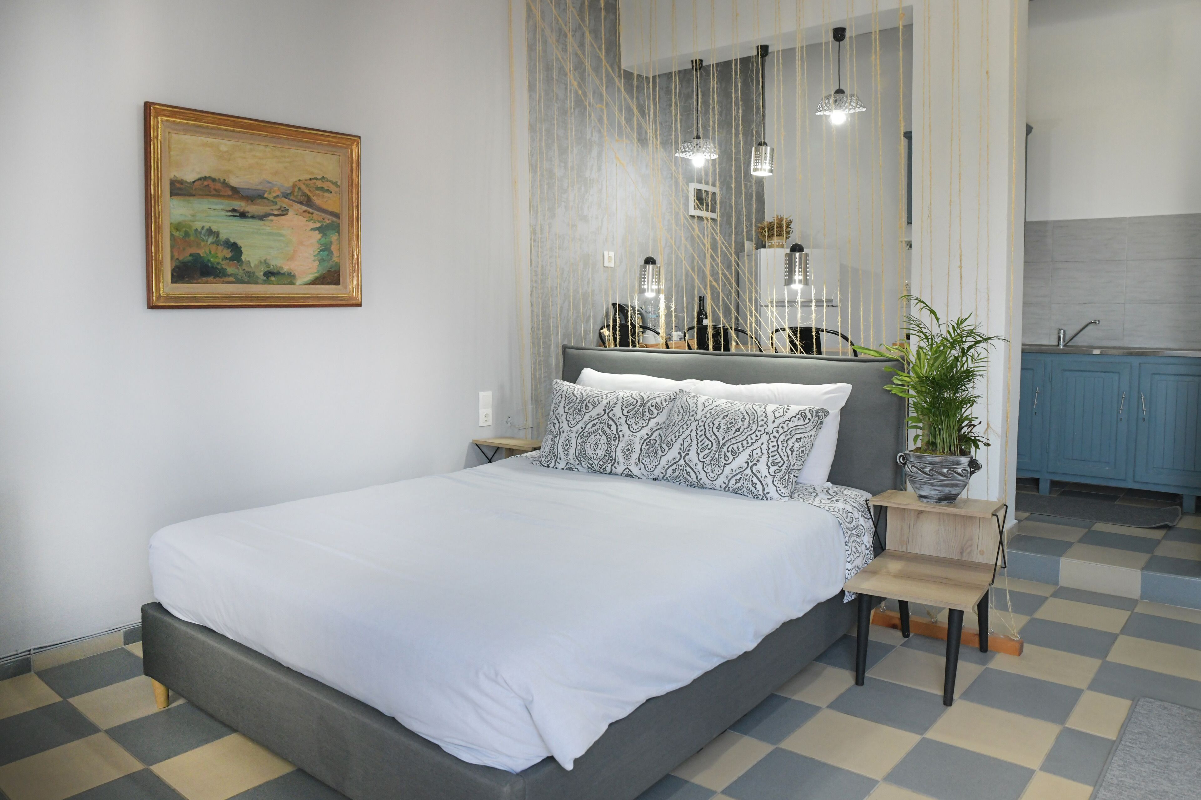 A modern bedroom with a large bed, a bedside table with a plant, a mirror, and a view of the kitchen.
