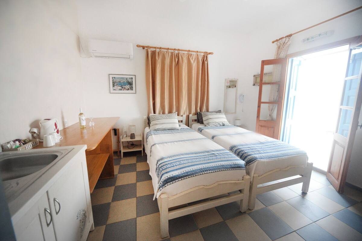 A room with two beds, a wooden desk, a sink, and a window with curtains.