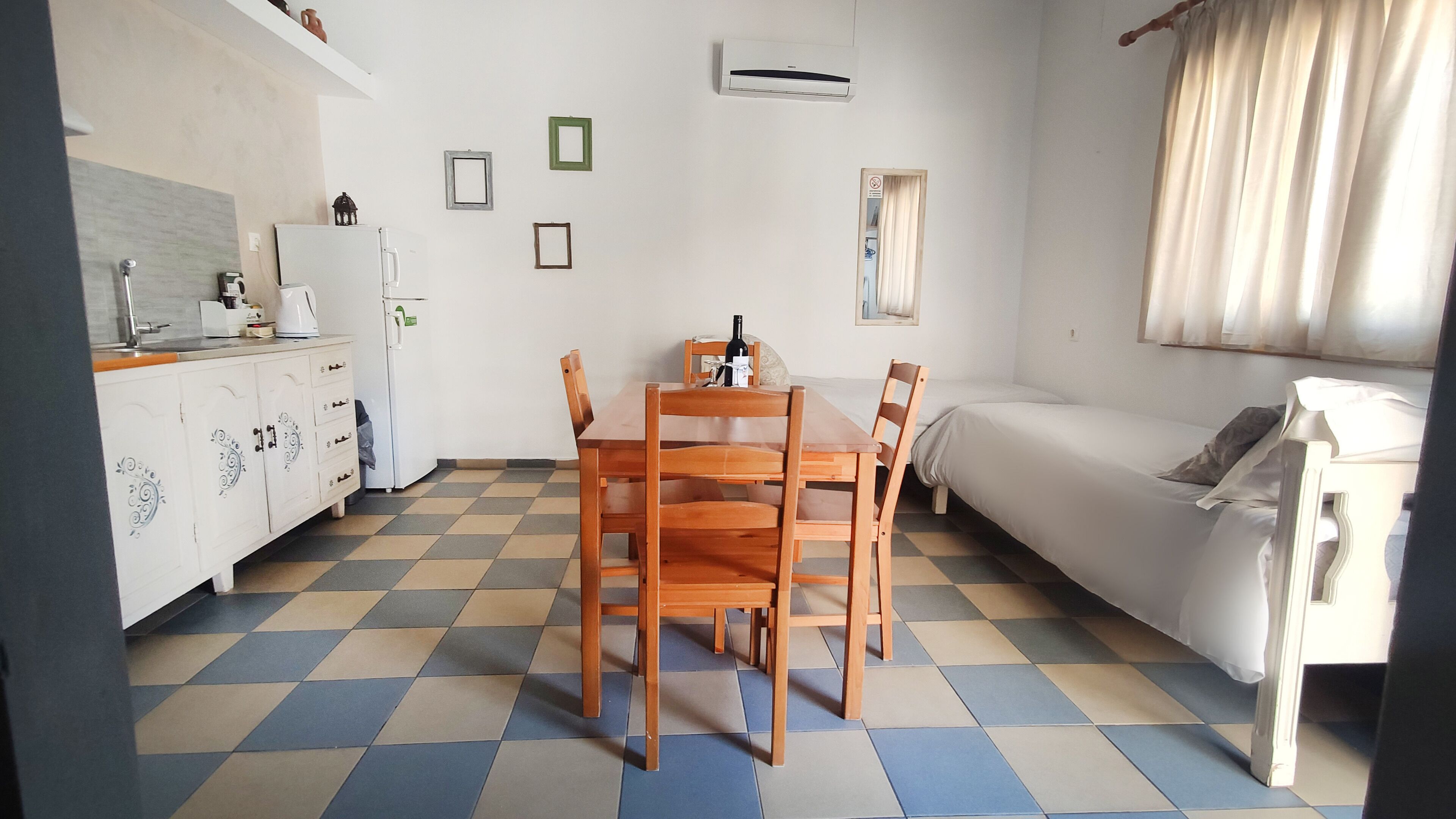 Ein kleines, sauberes Zimmer mit kariertem Boden, einem weißen Sofa, einem Holzespeisetisch mit Stühlen, einem weißen Schrank und einer Wand-Klimaanlage.
