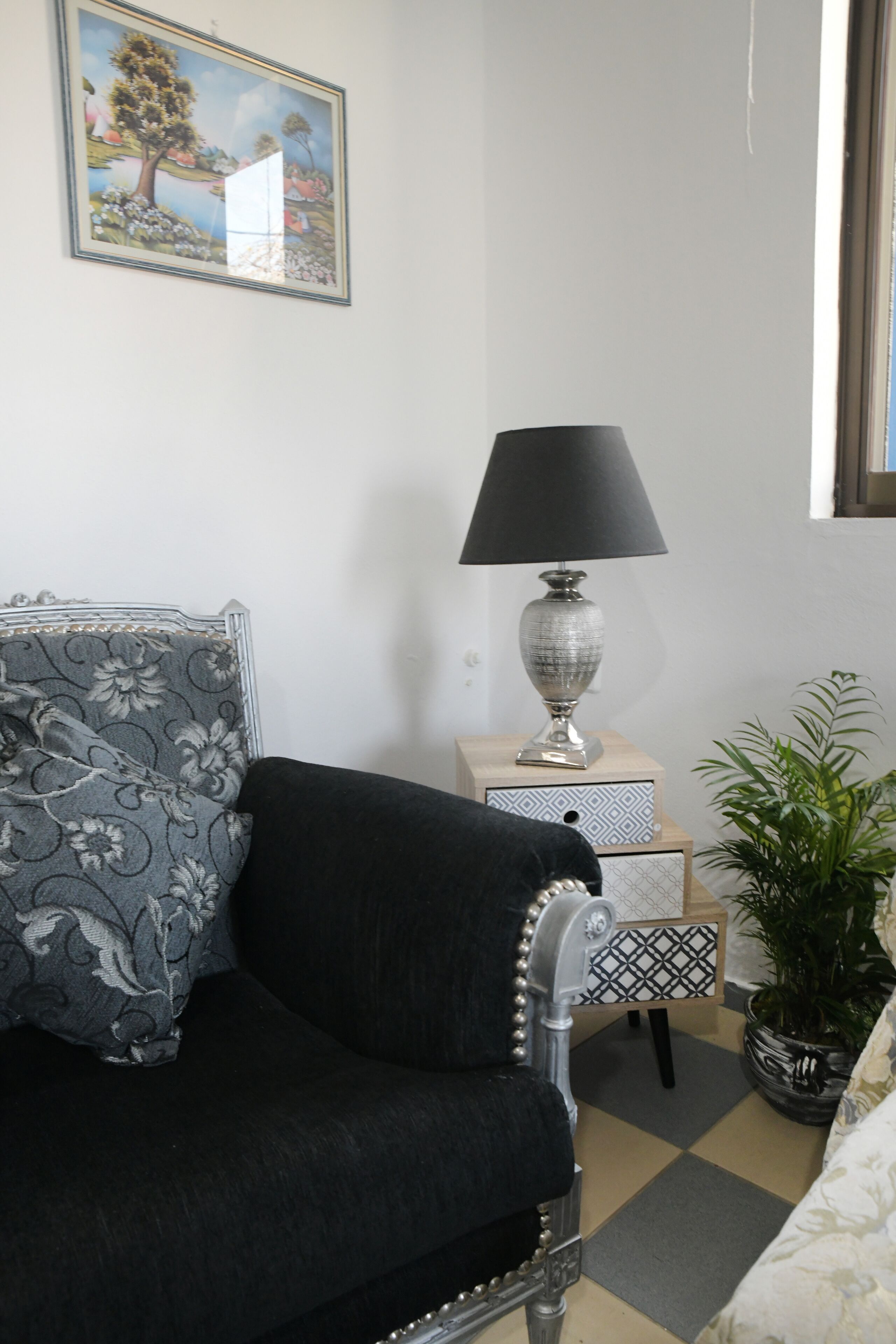 A room with a black sofa, a patterned pillow, a lamp on a side table, a potted plant, and a painting on the wall.