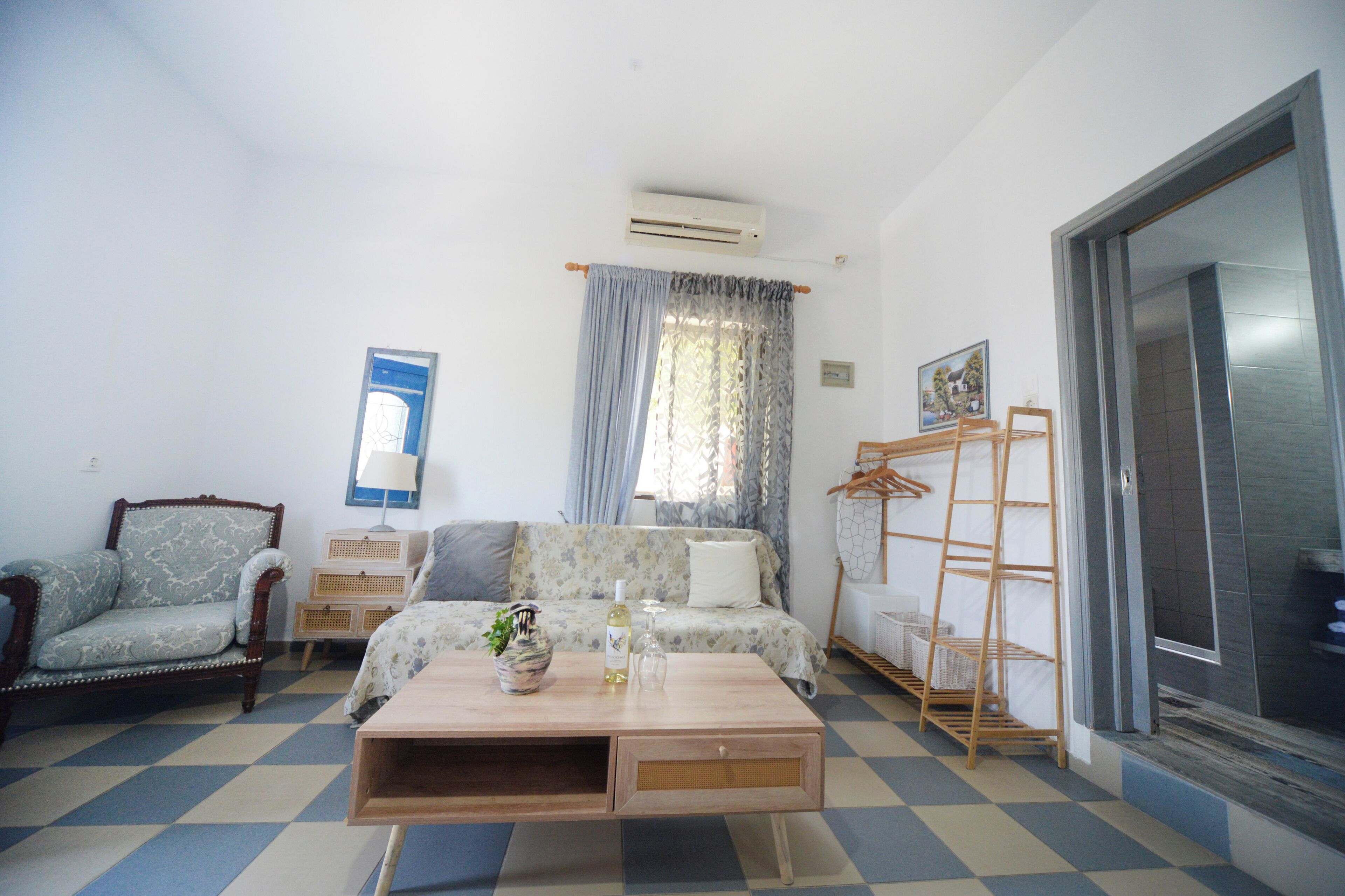 Una habitación con suelo a cuadros, un sofá, una silla, una mesa de centro de madera, un aire acondicionado, una ventana con cortinas y una puerta con perchero.