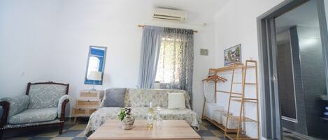 Una habitación con suelo a cuadros, un sofá, una silla, una mesa de centro de madera, un aire acondicionado, una ventana con cortinas y una puerta con perchero.