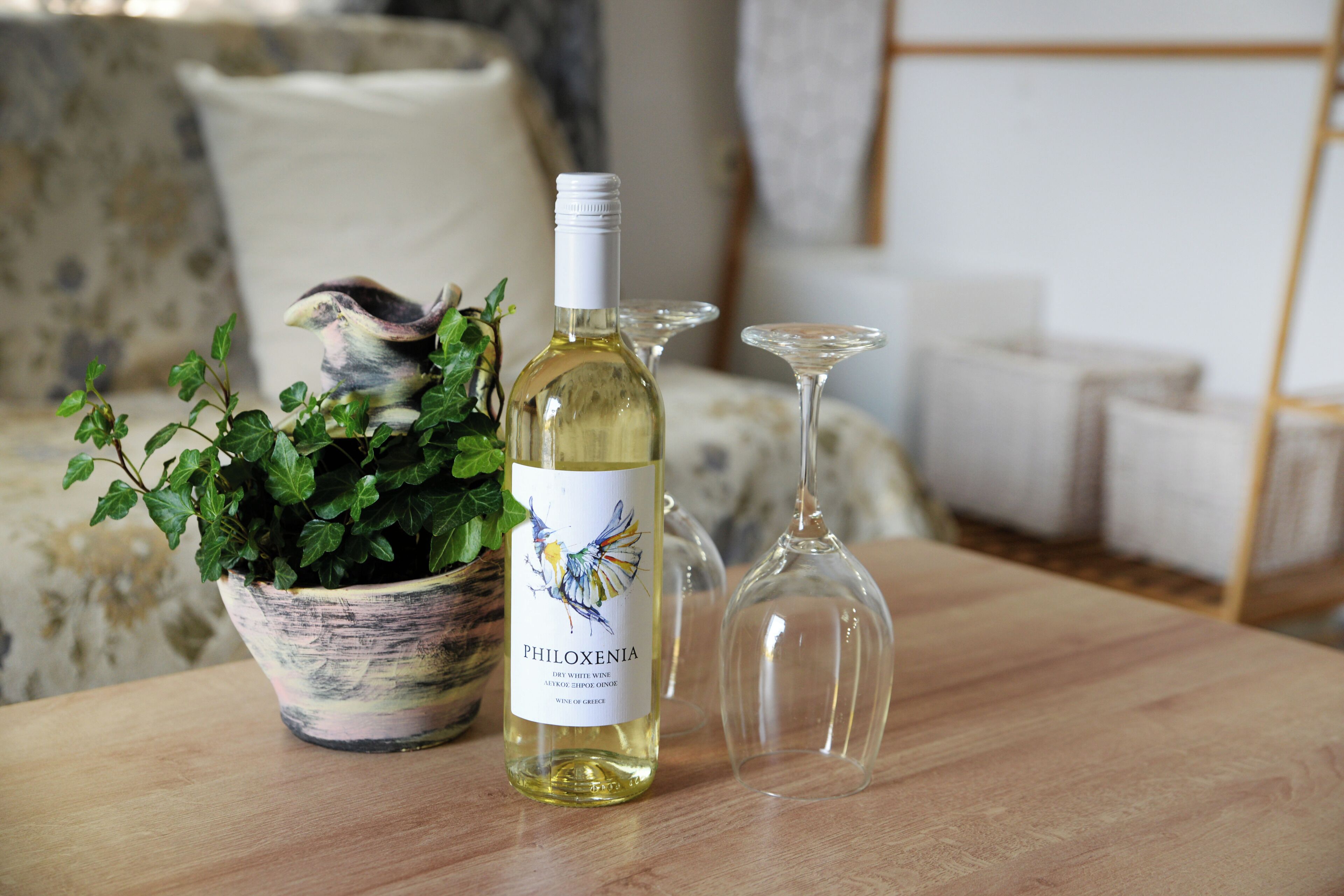 Una botella de vino Philoxenia, una planta en maceta y dos copas de vino sobre una mesa de madera.