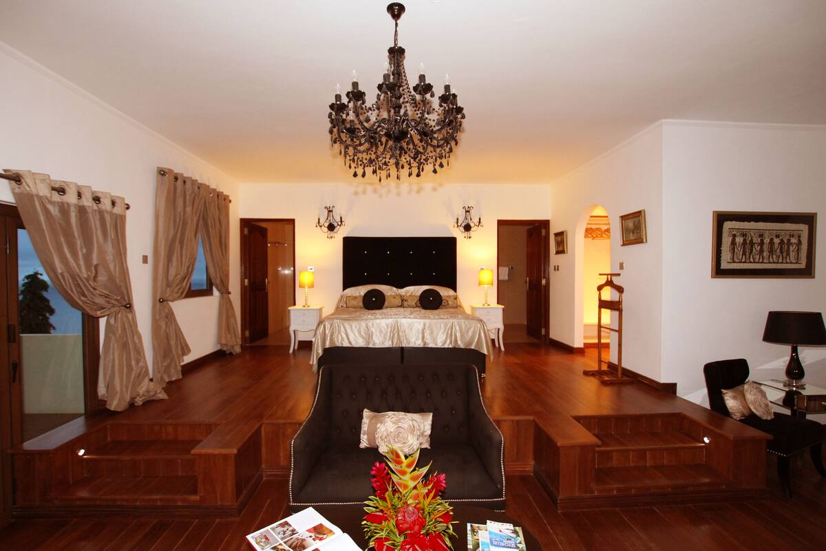 A hotel room with a large bed, a dark armchair, a coffee table with magazines, a chandelier, and a framed picture on the wall.