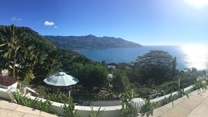 Una terraza con vistas al mar, rodeada de vegetación tropical y una montaña al fondo.