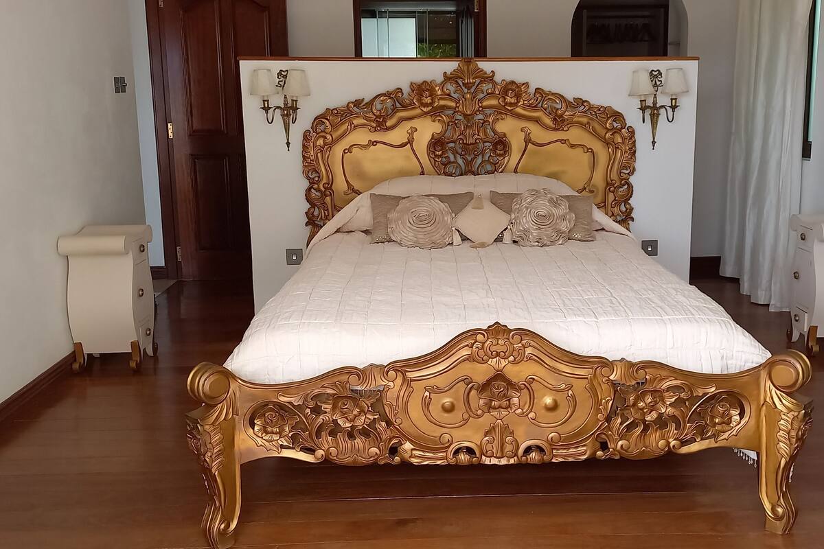 A large, ornate bed with a gold frame and white bedding, flanked by two nightstands and a white dresser with a mirror.