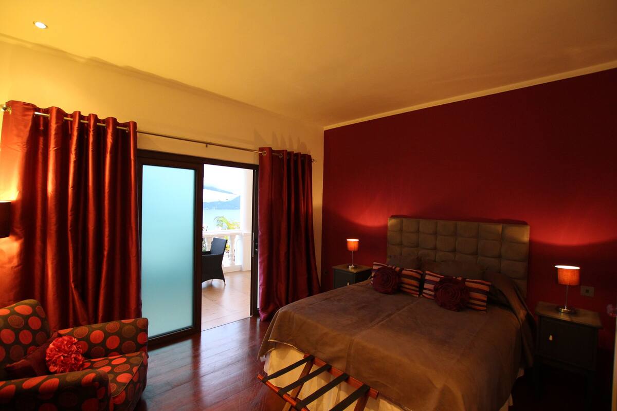 A bedroom with a large bed, a chair, a nightstand, and a view of the outdoors.