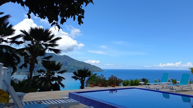 Zona de piscina con sillas de descanso y plantas en macetas, con vistas a un paisaje marino y montañas al fondo.