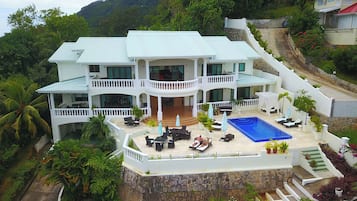 A large, multi-story house with a swimming pool, multiple balconies, and a stone retaining wall.