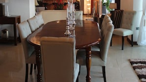 Un comedor con una mesa redonda, seis sillas y una araña. Se observa una cocina al fondo con un ventanal grande.
