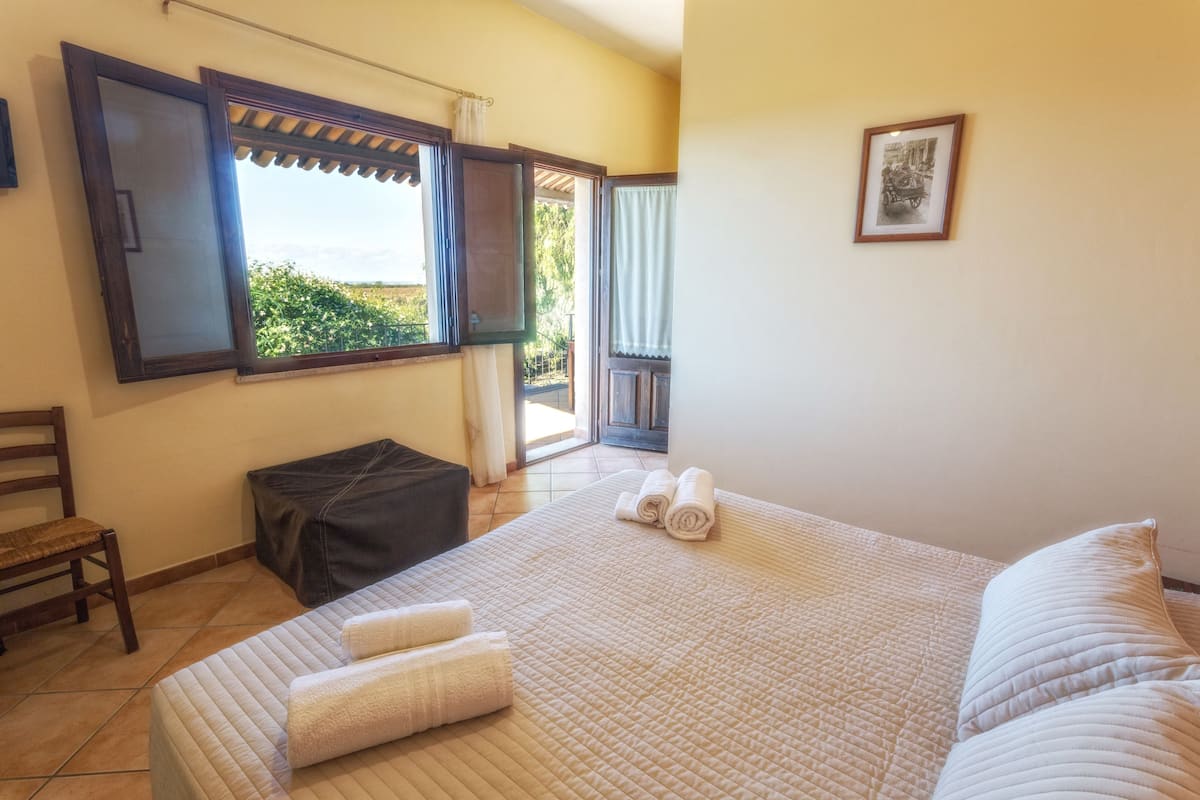 A bedroom with a large bed, a chair, a bench, and a window with a view of greenery.
