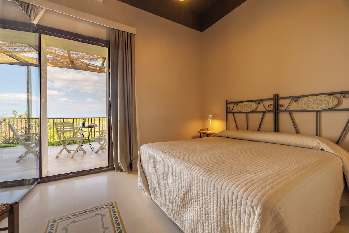 A bedroom with a large bed, a balcony with a table and chairs, and a view of greenery outside.