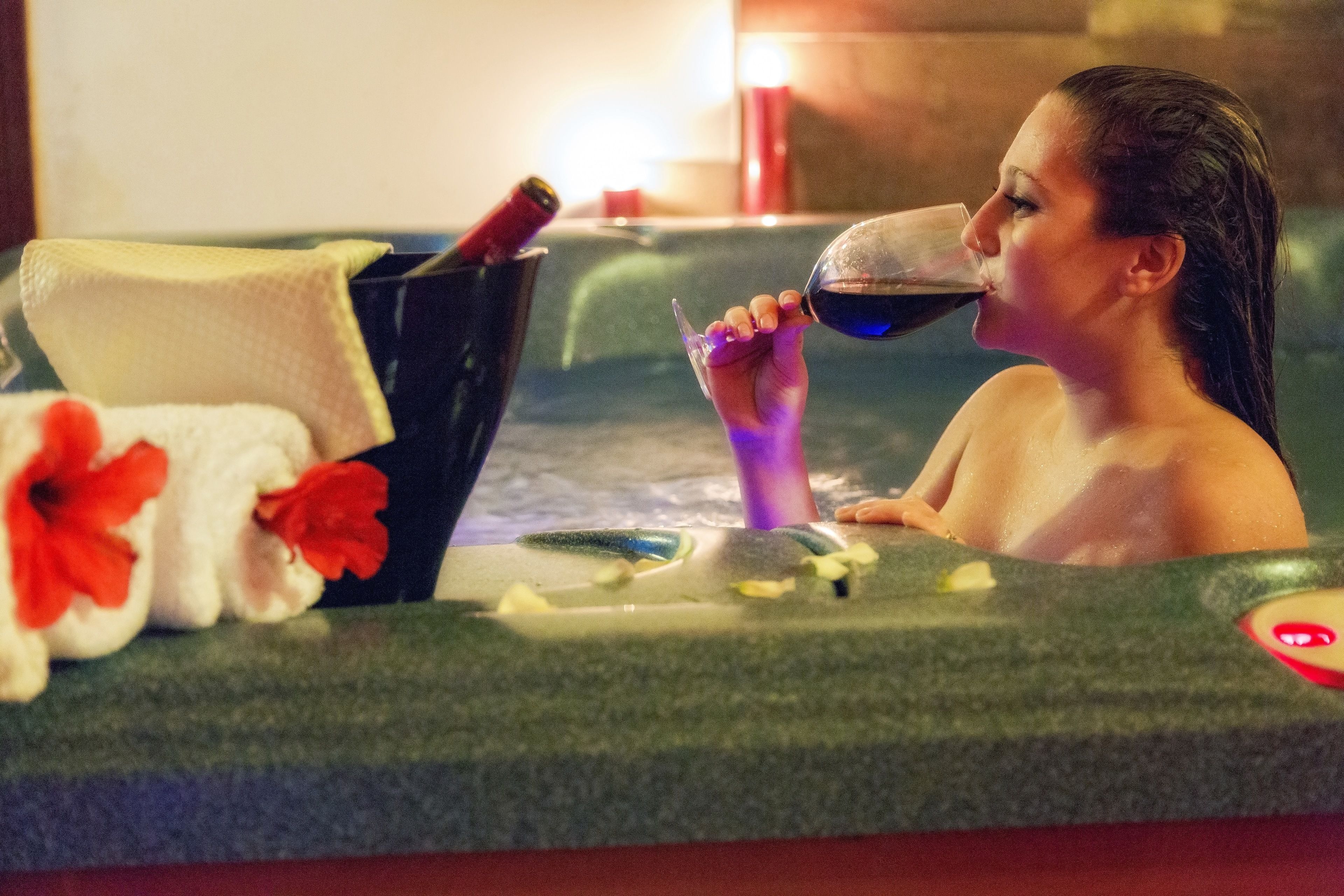 A person in a hot tub with a bottle and glass, a towel, and flowers.