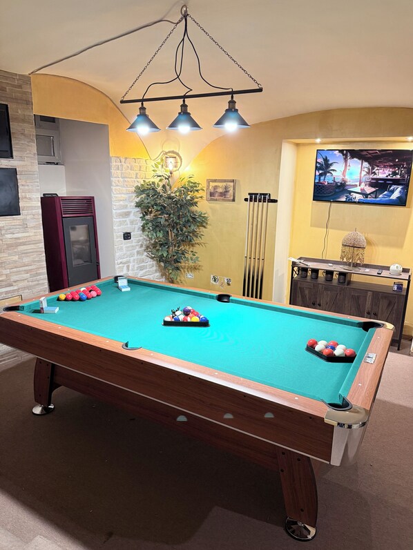 Una mesa de billar con bolas, un televisor y una planta en una habitación con un mueble de madera y una pared de piedra.