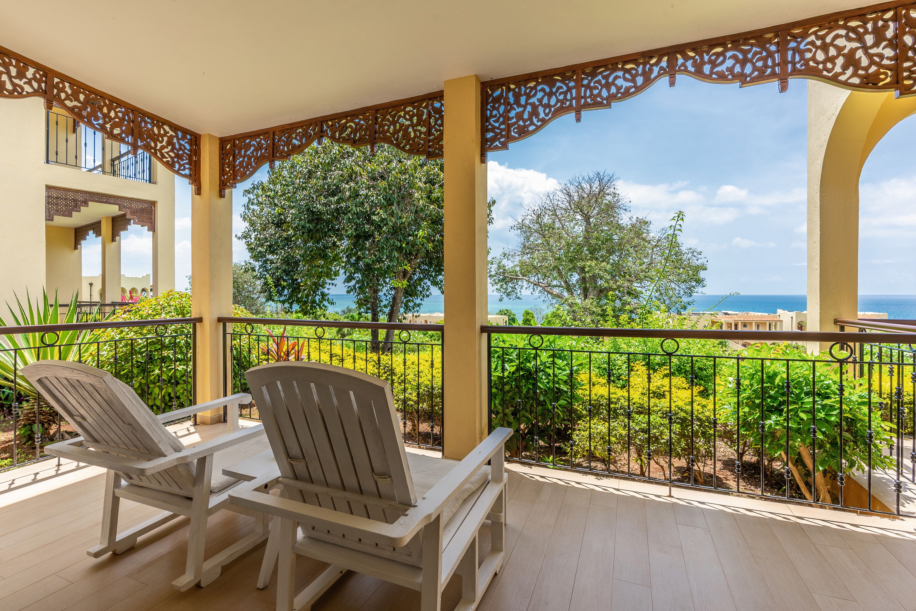 Un balcón con dos sillas Adirondack, una mesa de madera y vistas a un jardín ajardinado y el océano.