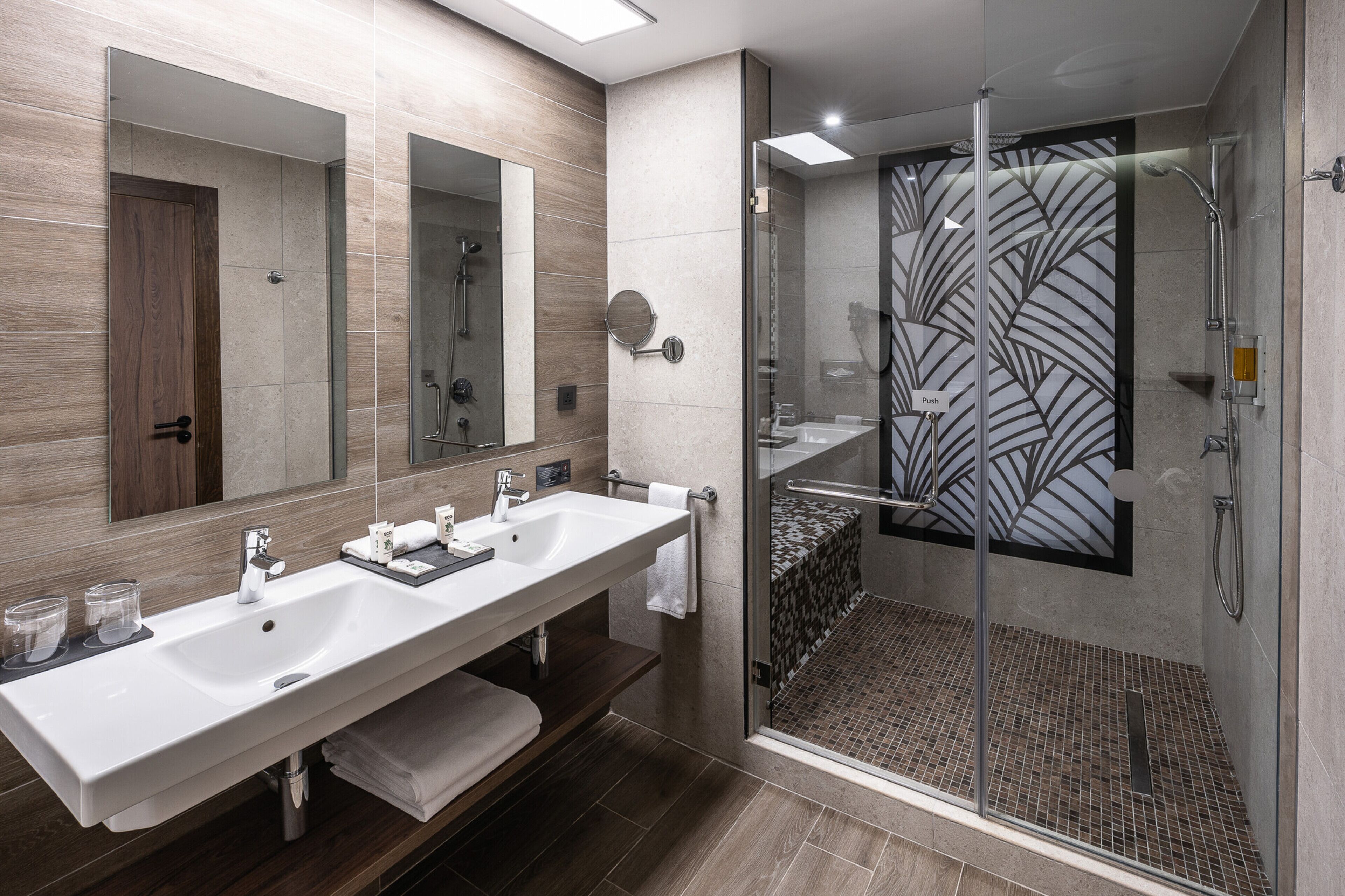 Un baño moderno con un lavamanos grande blanco, dos espejos, una ducha con mampara de vidrio y una pared con azulejos estampados.