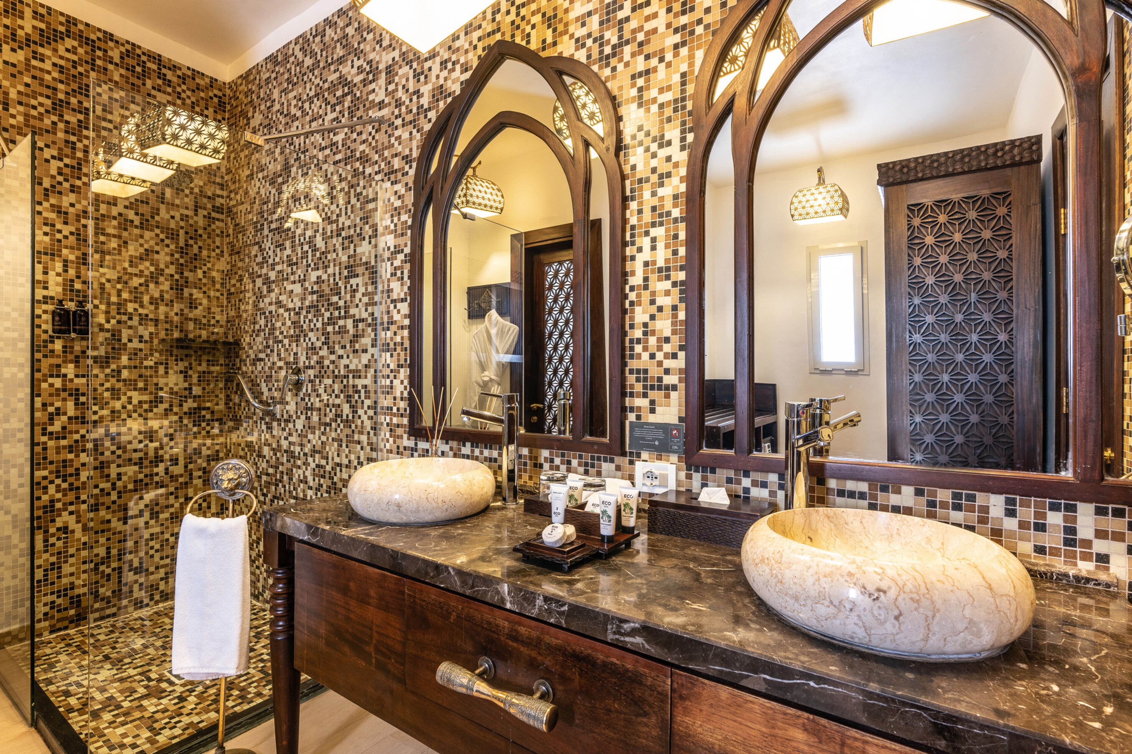 A bathroom with mosaic tile walls, two large oval sinks, and a mirror with an arched frame.