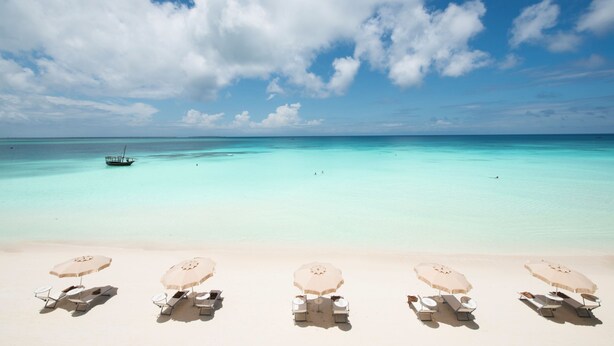 A beach with clear turquoise water, a boat in the distance, and several beach umbrellas and lounge chairs. - Hotel Riu Palace Zanzibar - All Inclusive (Nungwi)