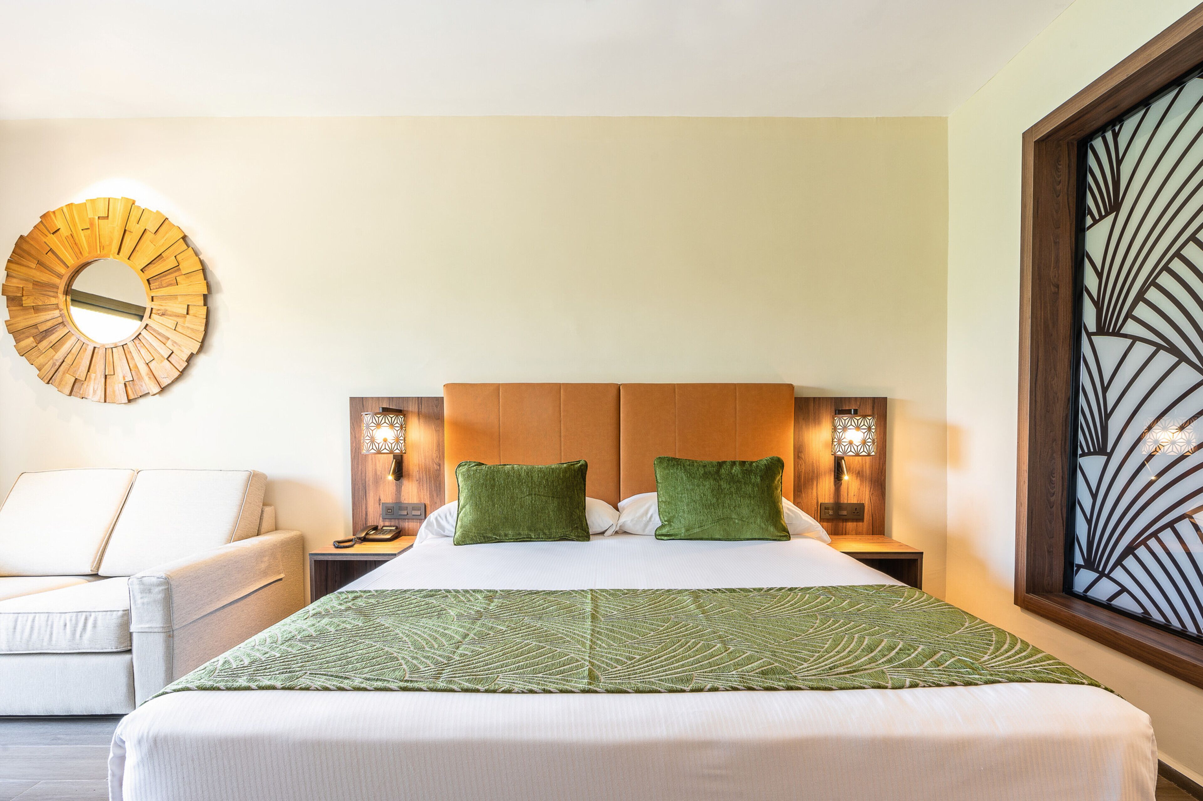 Habitación de hotel ordenada con una cama grande, dos almohadas verdes, un espejo redondo en la pared y una vista decorativa desde la ventana.