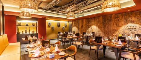 Een restaurant met houten tafels en stoelen, hangende rieten lampen en decoratieve wandkunst.