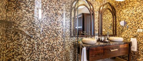 Un baño con paredes de mosaico, cabina de ducha de vidrio y tocador de madera con dos lavamanos.