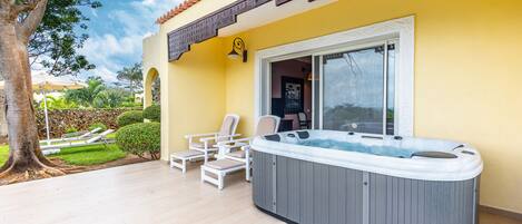 A hot tub in a sunlit outdoor area with lounge chairs and a view of a garden.