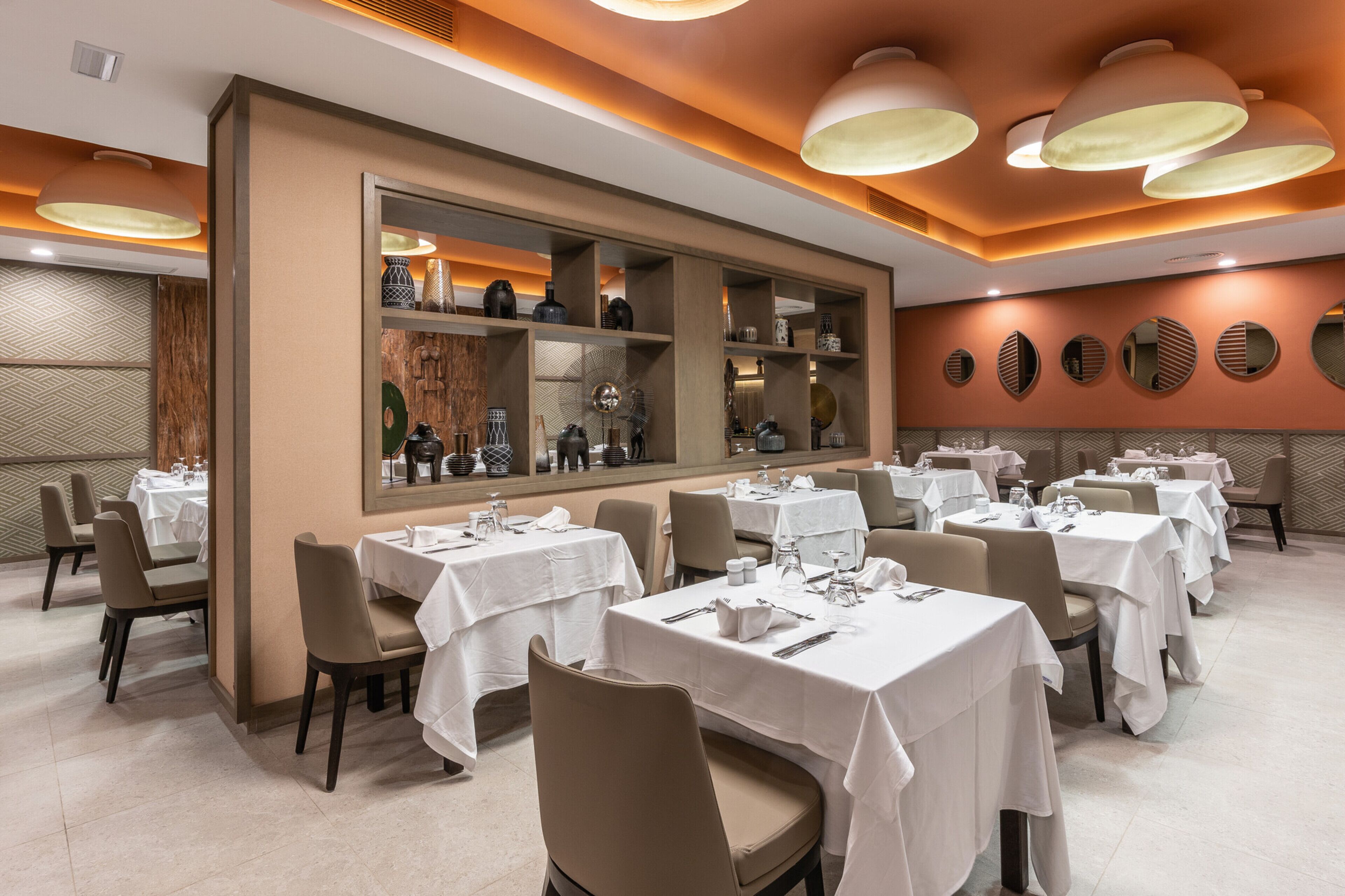 A restaurant with multiple tables, white tablecloths, and beige chairs.