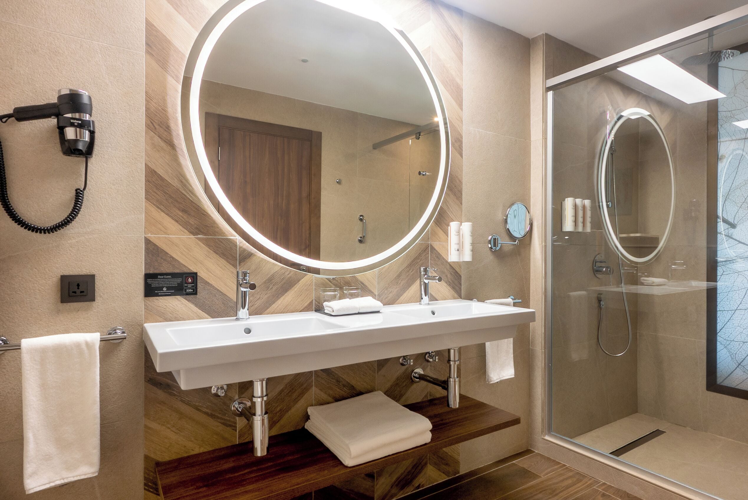 Une salle de bain moderne dotée d’un grand miroir, de deux vasques, d’une douche à parois de verre et d’un sèche-cheveux fixé au mur.