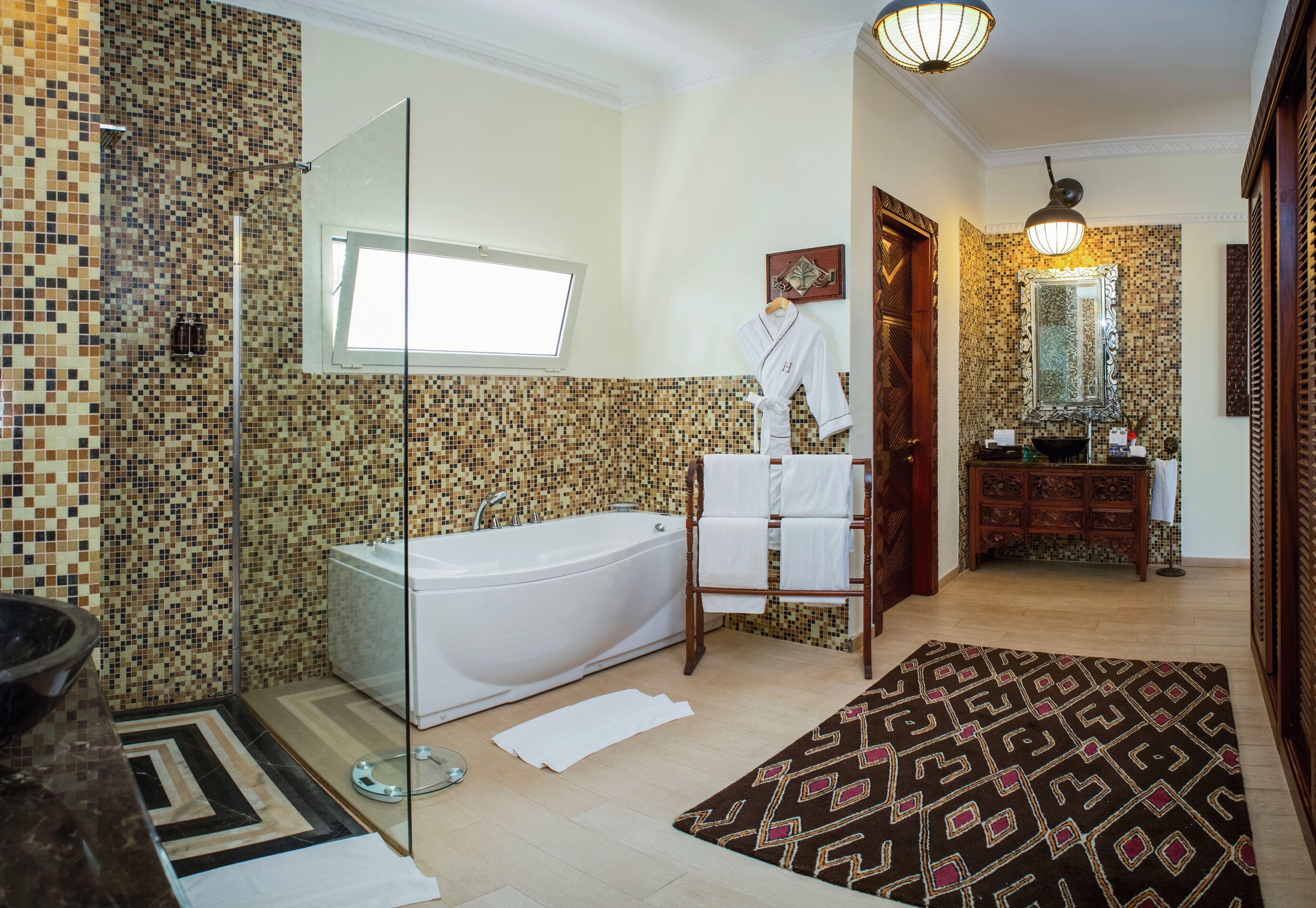 Un baño con pared de mosaicos, una bañera, cabina de ducha de vidrio, tocador de madera con espejo y alfombra estampada en el suelo.