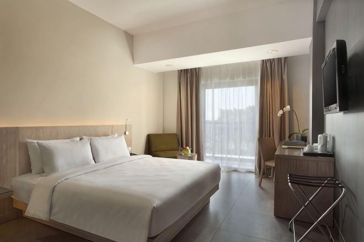 Una habitación de hotel con cama, escritorio, silla, televisor y ventana con cortinas.