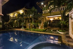 Un área de piscina con una superficie de agua azul cristalina, rodeada de exuberante vegetación y un edificio bien iluminado con múltiples balcones y un espacio de bar.