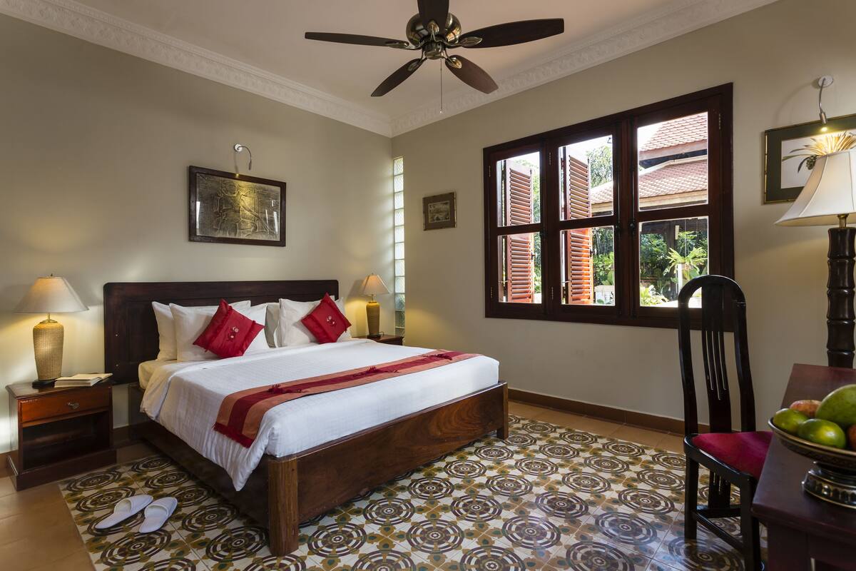 Una habitación de hotel con una cama grande, un ventilador de techo, una ventana con contraventanas, una silla y una mesa con fruta.