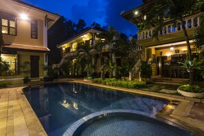 A swimming pool with a tiled edge, surrounded by a landscaped garden with palm trees and a building with a balcony. - HanumanAlaya Colonial House (Siem Reap)