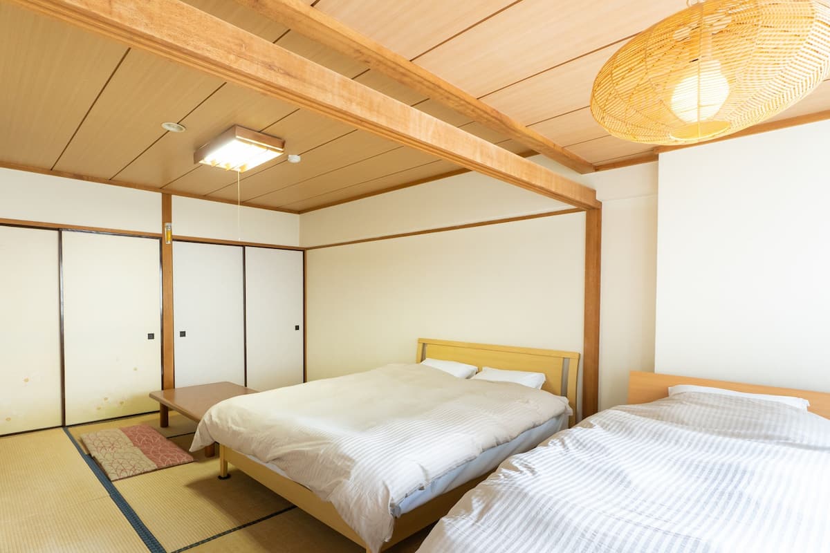 A traditional Japanese-style room with tatami flooring, two wooden beds, a ceiling fan, and a woven light fixture.