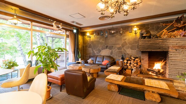 A cozy lounge area with a stone fireplace, comfortable seating, and a large window offering a view of the outdoors. - Villa Alpen (Yamanouchi)