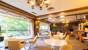 A modern dining area with round tables and chairs, a stone fireplace, and large windows offering a view of trees. - Villa Alpen (Yamanouchi)