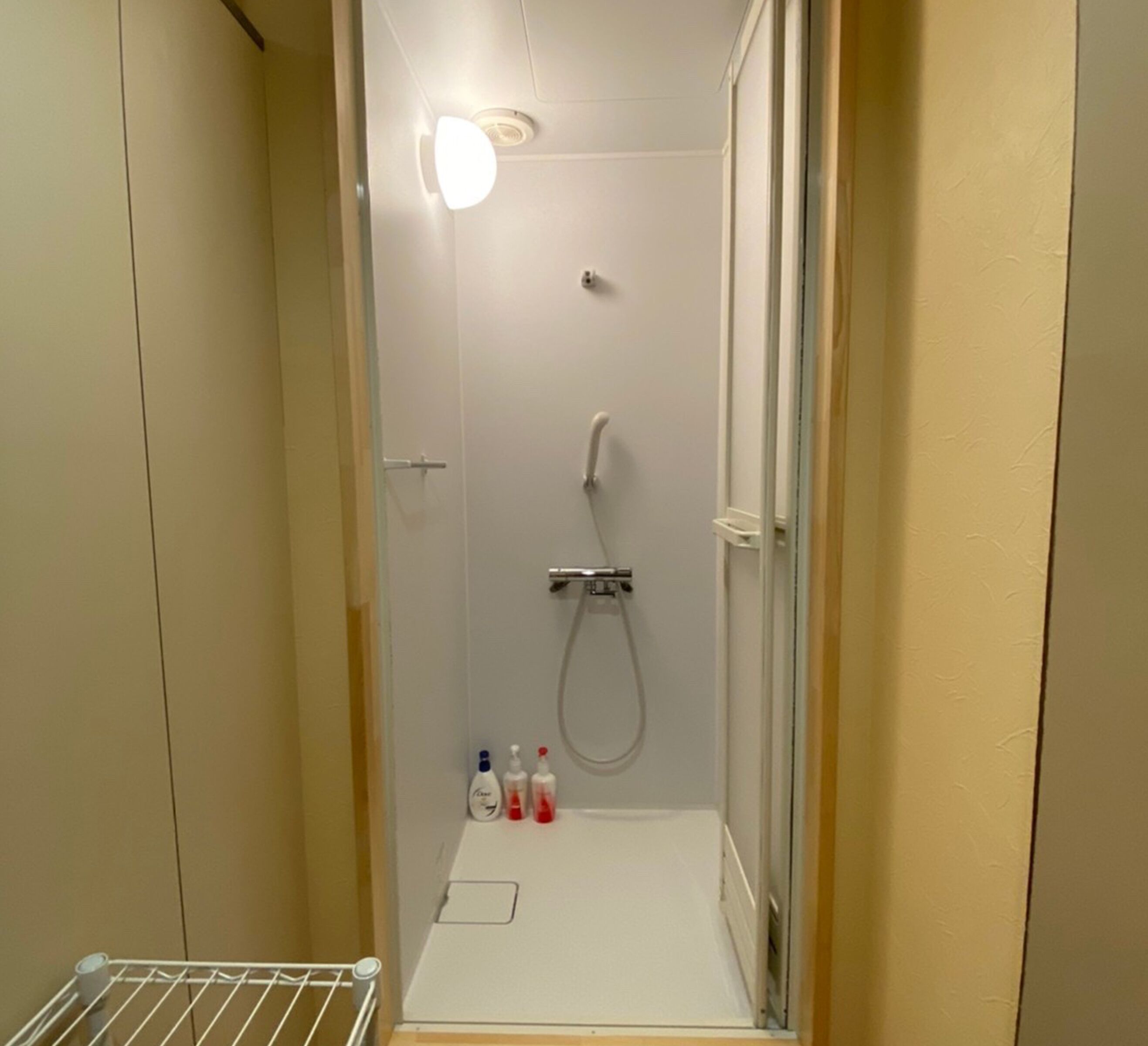 Un petit espace douche fermé, avec un sol blanc, une douchette et un robinet fixé au mur. On y trouve trois bouteilles de savon liquide sur le sol.