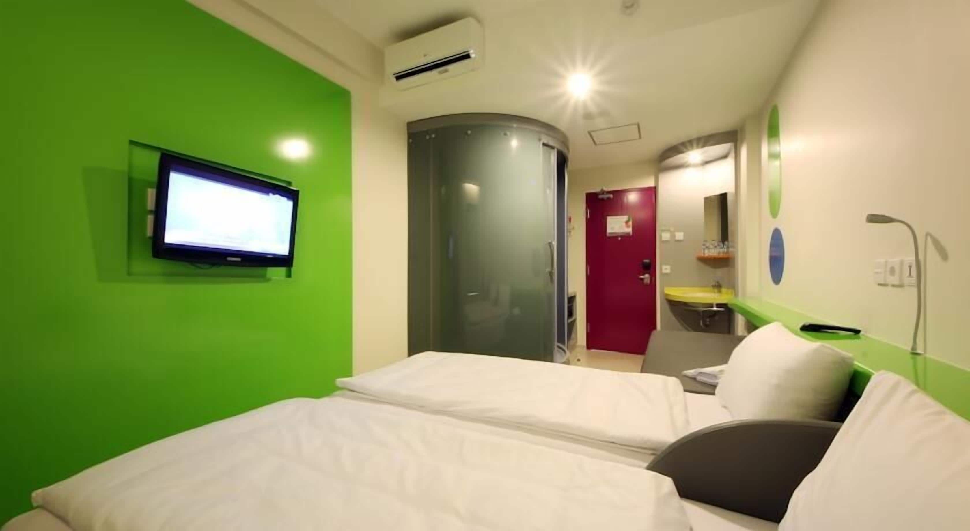 Habitación de hotel con pared verde, televisor de pantalla plana, cama, zona de ducha y baño con lavamanos y espejo.