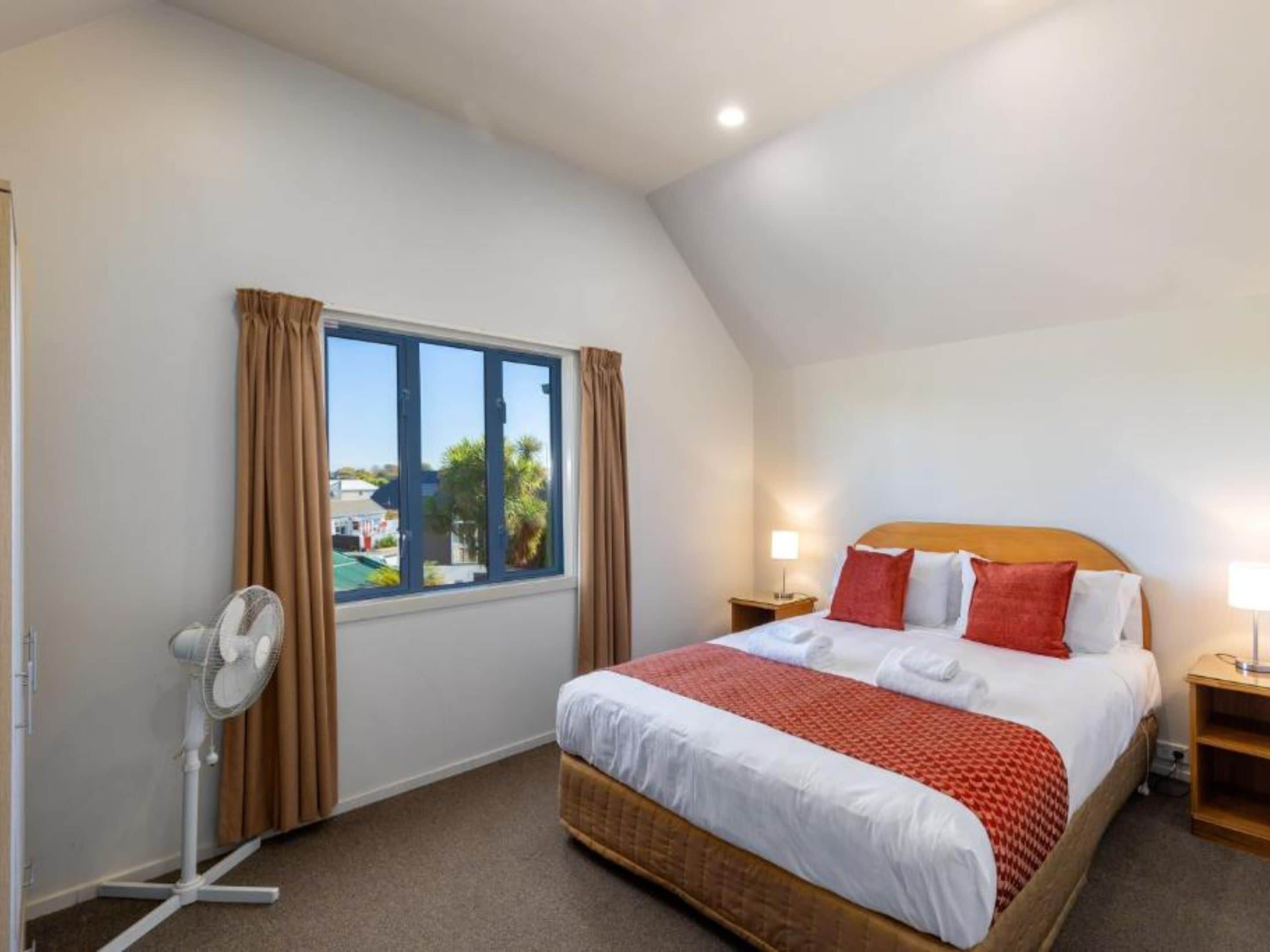 Un dormitorio con una cama grande, un ventilador de pie, una mesita de noche con lámpara y una ventana con cortinas.