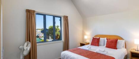 A bedroom with a large bed, a standing fan, a nightstand with a lamp, and a window with curtains.